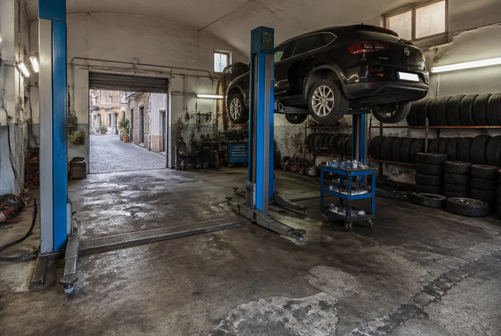 L'officina vista da dentro: due ponti sollevatori, un'auto in alto sul secondo ponte, le gomme accatastate lungo la parete di destra e la porta a rullo aperta sul vicolo.