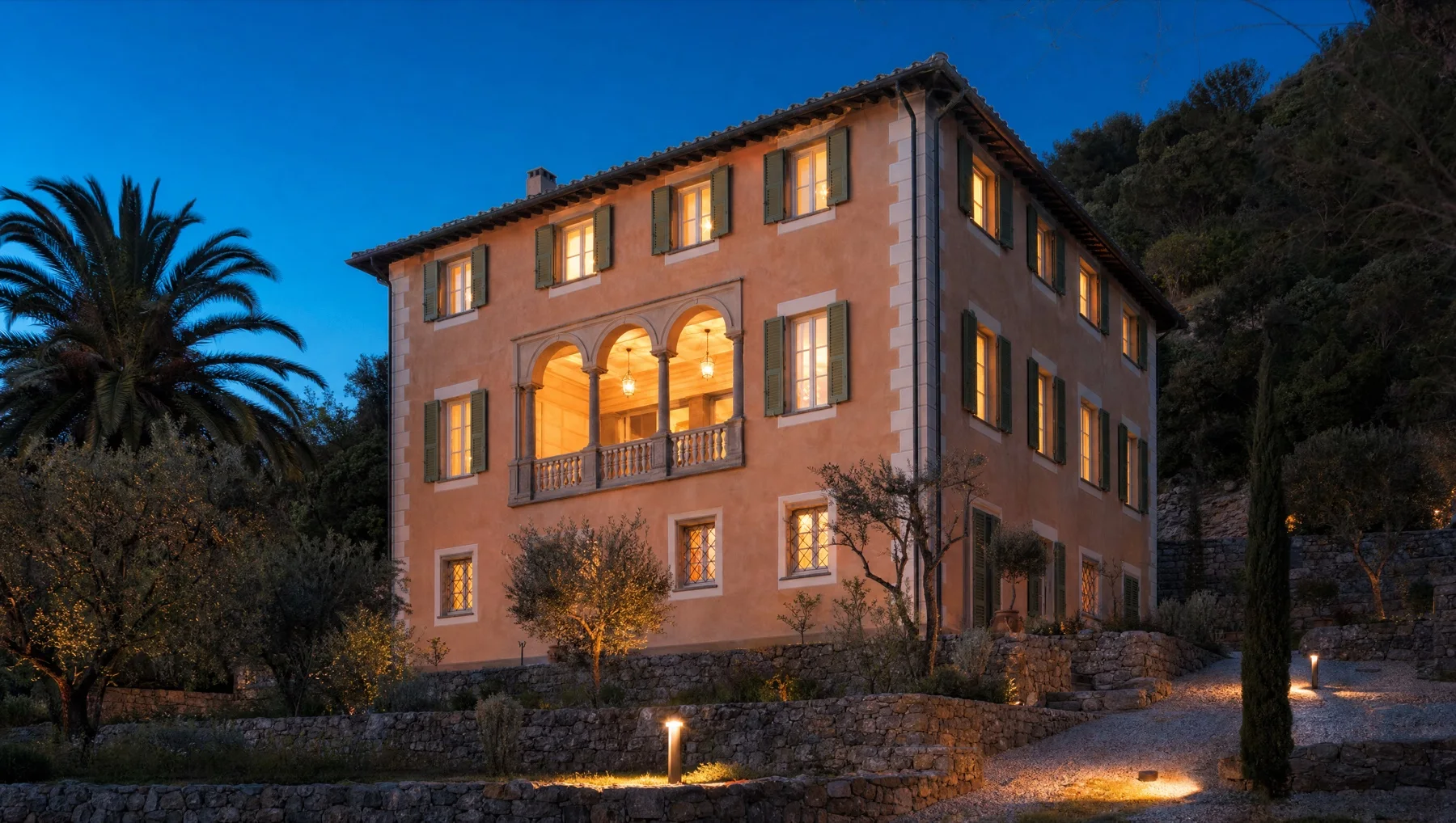 Villa ottocentesca intonacata di rosa antico con persiane verdi e loggia a tre archi, illuminata dall'interno all'ora blu, fra ulivi e muri a secco