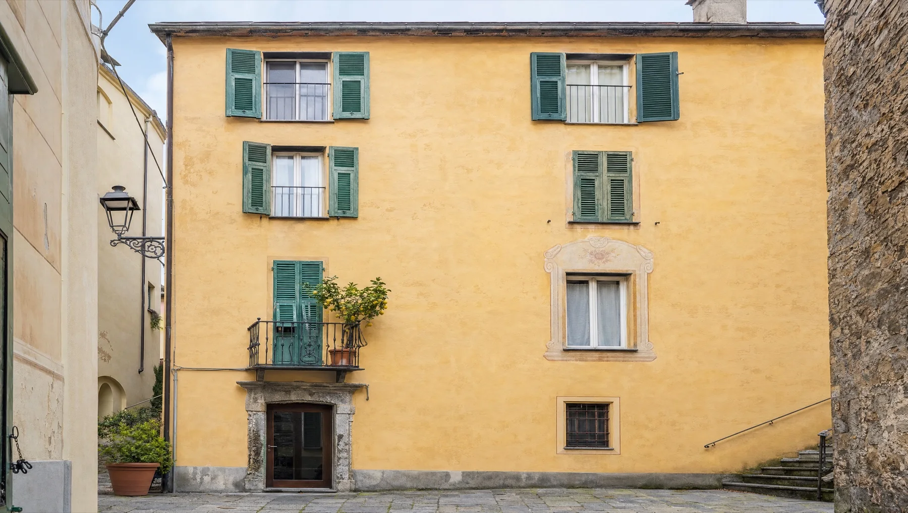 Facciata gialla di una casa di paese con persiane verdi, un balconcino in ferro con un limone in vaso e la piazzetta lastricata davanti