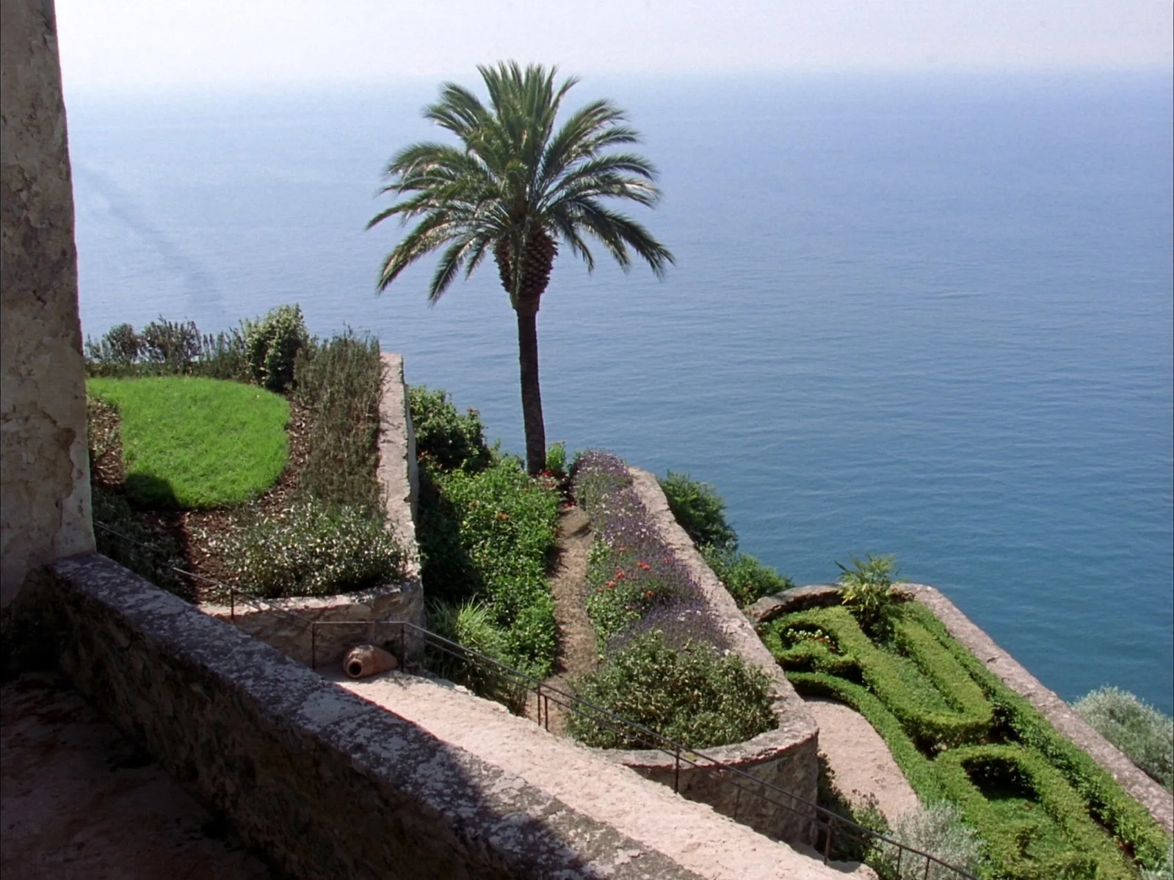 Terrazza del giardino con muri a secco, aiuole di bosso potato e una palma alta, il mare aperto che occupa la metà del quadro