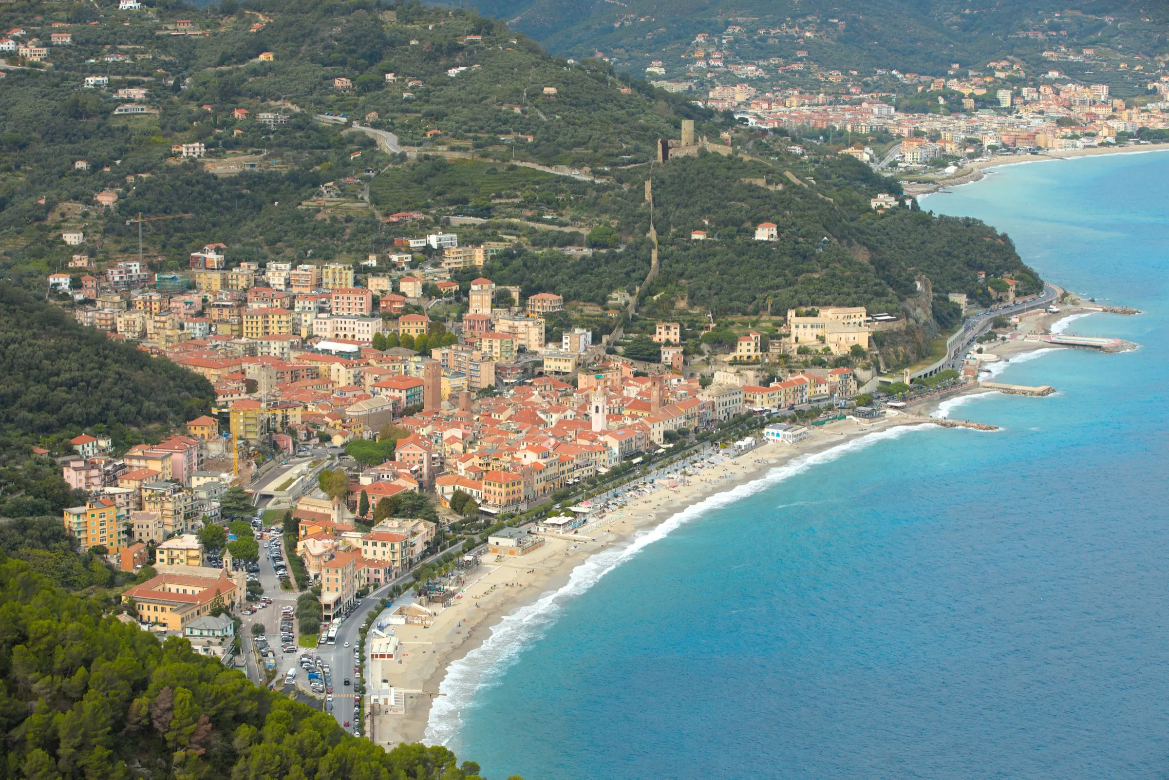 Veduta aerea di un paese di costa: il centro storico di case addossate, la spiaggia lunga, la strada che gira dietro il capo e le fasce di ulivi sulla collina